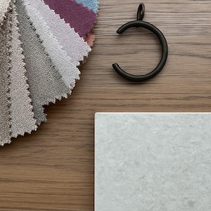 May include: A collection of fabric swatches in various colors, including beige, pink, and blue, alongside a dark metal curtain ring and a square, light gray tile. The items are arranged on a wooden surface.