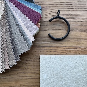 May include: A collection of fabric swatches in various shades of gray, beige, brown, pink, blue, and black. The swatches are arranged in a fan shape and are displayed on a wooden surface. A black metal curtain ring is also visible in the image.