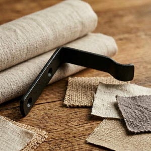 May include: A black metal bracket with two holes, angled against a backdrop of rolled beige fabric and fabric swatches. The swatches are in various neutral tones, including beige and brown, on a wooden surface.