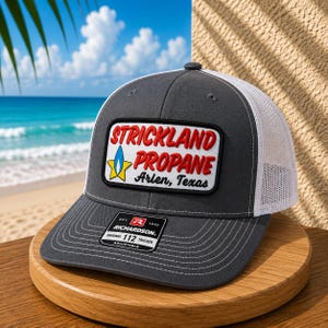May include: A charcoal gray and white trucker hat featuring a rectangular patch with the red text Strickland Propane and a blue and yellow flame icon. The patch also includes the black text Arlen, Texas. A black Richardson 112 sticker is on the brim.