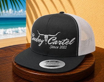 May include: A black snapback trucker hat with a white mesh back. The front features white embroidery of a longhorn skull icon above the text Cowboy Cartel Since 2021. A circular sticker on the flat brim reads The Authentic Yupoong Snapback.