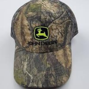 May include: A camouflage baseball cap with a green John Deere logo featuring a leaping deer. The logo is outlined in black and the text "John Deere" is in black below the logo.