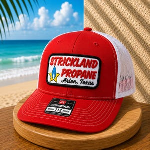 May include: A red and white trucker hat featuring a rectangular patch with the text Strickland Propane Arlen, Texas and a flame graphic. The front panel is solid red, while the back panels are white mesh. A black sticker is on the brim.
