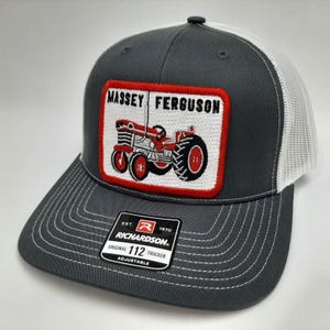 May include: A gray and white trucker hat with a red and white patch featuring a red Massey Ferguson tractor. The patch has the text "Massey Ferguson" in white. The hat has a black and red Richardson label with the text "EST. 1970 ORIGINAL 112 TRUCKER ADJUSTABLE".