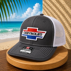 May include: A charcoal gray trucker hat with a white mesh back and an adjustable snapback closure. The front features an embroidered Chevrolet logo patch in red, white, and blue. A black Richardson 112 sticker is affixed to the curved brim.
