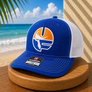 May include: A royal blue and white trucker hat featuring a white mesh back and a front panel with an embroidered circular logo. The logo displays an orange sun and a white alligator silhouette above a white Florida state outline. A black sticker is on the brim.