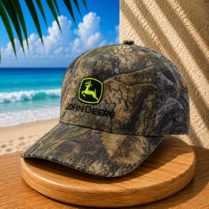 May include: A camouflage baseball cap featuring a brown, green, and black leaf pattern. The front panel displays a bright lime green embroidered John Deere logo with a leaping deer icon and the text John Deere in black lettering.