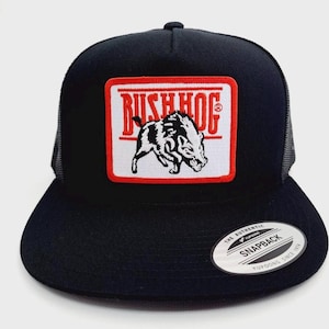 May include: Black trucker hat with a flat brim and mesh back. The front features a rectangular patch with the text "BUSH HOG" in red and a black and white illustration of a boar. The hat has a snapback closure.