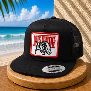 May include: A black trucker hat featuring a rectangular patch with a red border. The patch displays the text BUSH HOG in red letters above a black and white illustration of a wild boar. A circular silver sticker is on the flat black brim.