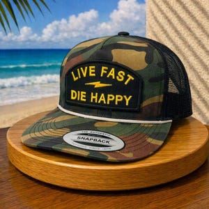 May include: A camouflage print snapback cap with a black mesh back and a black rectangular patch on the front. The patch features the yellow text LIVE FAST DIE HAPPY with a lightning bolt icon. A white cord trim separates the crown and brim.