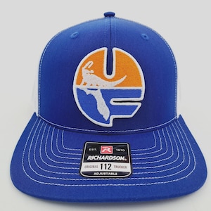 May include: A blue and white trucker hat with a curved brim. The hat features a patch with an orange and white design, including a stylized alligator and the outline of Florida. The hat has the text "RICHARDSON" and "112 TRUCKER".