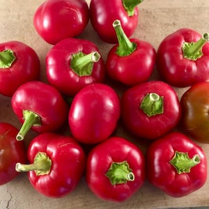 May include: A selection of bright red cherry peppers, each with a green stem and calyx. The peppers are displayed on a light brown surface, emphasizing their smooth, shiny skin. The image showcases fresh, natural produce.