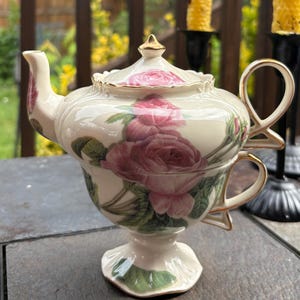 Puede incluir: Un juego de té de porcelana color crema para una persona, con una tetera apilada sobre una taza. El conjunto está decorado con rosas rosadas y hojas verdes, con detalles en oro en el remate de la tapa, el asa y el borde. La base es de pedestal.