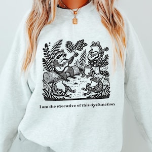 May include: A light grey crewneck sweatshirt featuring a black graphic print of a raccoon and three frogs playing musical instruments in a forest setting, with the text "I am the executive of this dysfunction" printed below the illustration.
