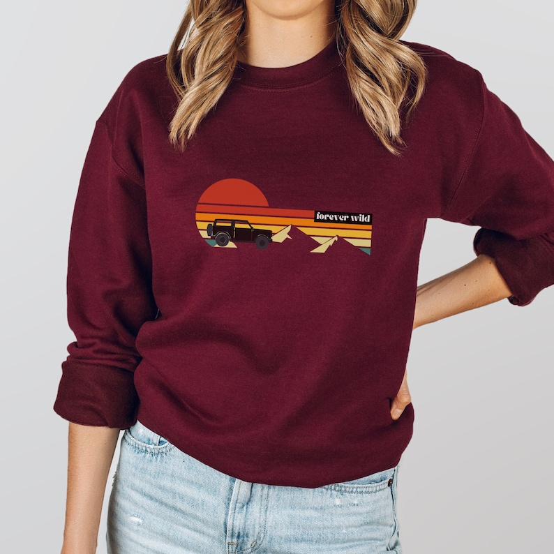 May include: A maroon sweatshirt with a retro sunset graphic featuring a jeep driving through mountains. The text "forever wild" is printed on the sweatshirt.