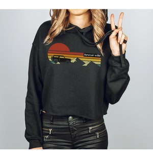 May include: Black cropped hoodie with a retro sunset graphic featuring a silhouette of a jeep driving through mountains. The text "forever wild" is printed on the graphic.