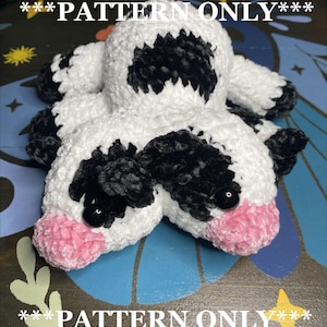 Peut inclure: Peluche de vache au crochet faite à la main en blanc, noir et rose. La peluche a des taches noires, des yeux en bouton et des sabots roses. Le texte "PATTERN ONLY" est écrit en blanc au-dessus et en dessous de la peluche.