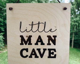 May include: A wooden sign with the text "little man cave" in black lettering. The sign is hanging from a brown rope and has a slightly pointed bottom.
