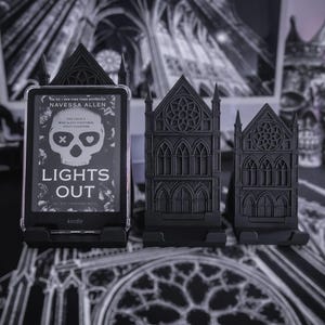 Könnte beinhalten: Schwarzer Kindle-Ständer im gotischen Stil, der ein Kindle-Gerät mit dem Buch "Lights Out" von Navessa Allen hält. Zwei zusätzliche schwarze Ständer in Form gotischer Gebäude sind ebenfalls vorhanden. Der Hintergrund zeigt ein gotisches Kathedralen-Design.