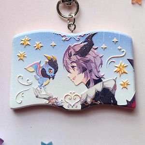May include: A rectangular keychain featuring a colorful anime-style illustration of a character with purple hair and a small blue dragon. The keychain has a silver heart-shaped clasp and is surrounded by paper stars and flowers.