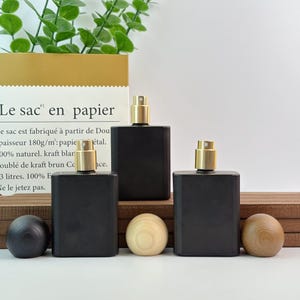 May include: Three black rectangular perfume bottles with gold-colored spray tops are displayed. The bottles are arranged in a row, with two wooden spheres and one dark sphere in front. A paper bag with French text is in the background.