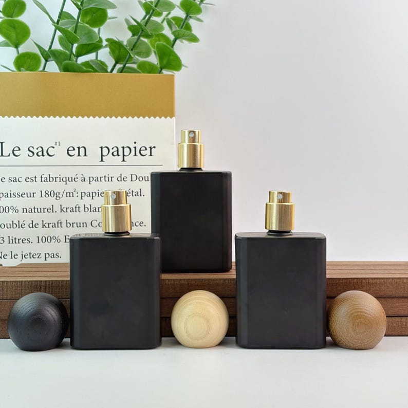 May include: Three black rectangular perfume bottles with gold-colored spray tops are displayed. The bottles are arranged in a row, with two wooden spheres and one dark sphere in front. A paper bag with French text is in the background.