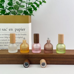 May include: Five small, round, frosted glass roller bottles with metal roller balls and wooden caps. The bottles are colored in clear, amber, pink, brown, and light green. Two additional wooden caps are placed in front of the bottles.