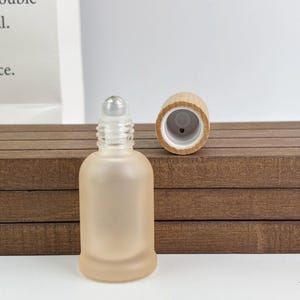 May include: A small, frosted glass roller bottle with a clear rollerball top and a separate wooden cap. The bottle has a rounded shape and a soft, peach colour. The wooden cap is light brown.