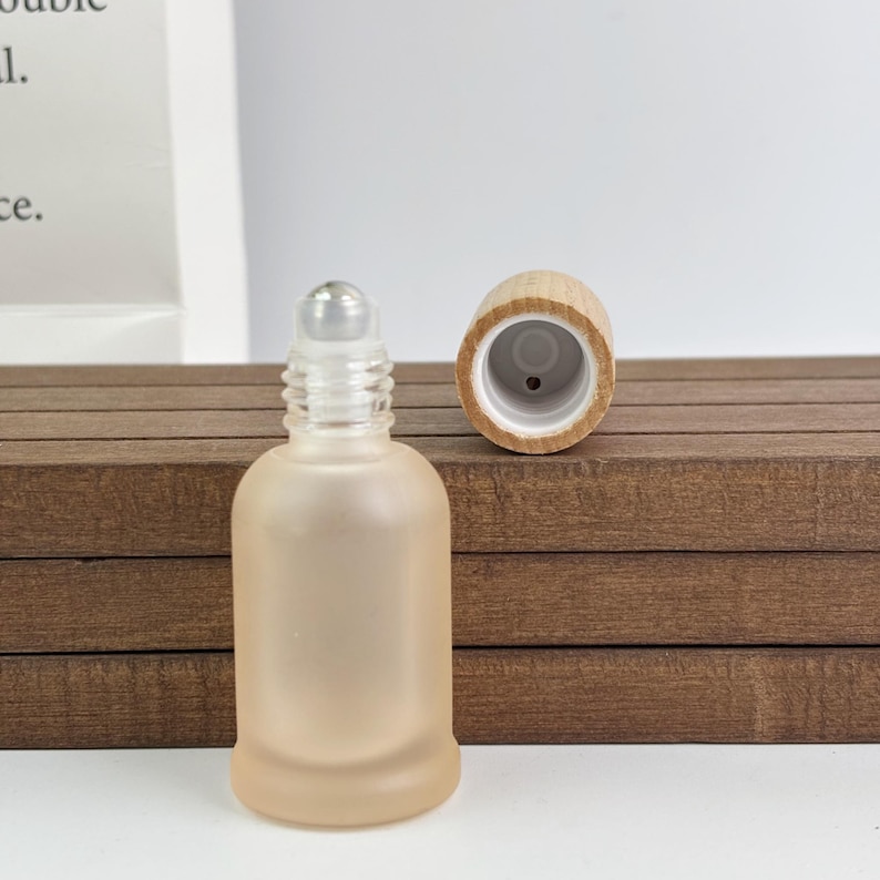 May include: A small, frosted glass roller bottle with a clear rollerball top and a separate wooden cap. The bottle has a rounded shape and a soft, peach colour. The wooden cap is light brown.