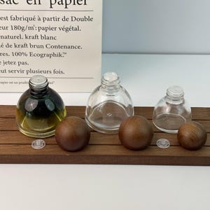 May include: Three glass bottles with wooden stoppers and clear plastic inserts are arranged on a dark wooden tray. One bottle contains a dark green and yellow liquid. The bottles vary in size and shape.