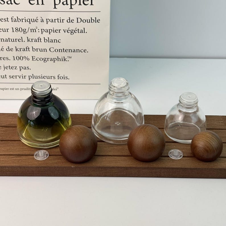 May include: Three glass bottles with wooden stoppers and clear plastic inserts are arranged on a dark wooden tray. One bottle contains a dark green and yellow liquid. The bottles vary in size and shape.
