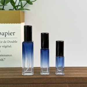 May include: Three glass roll-on perfume bottles with black caps. The bottles have a gradient blue color that fades to clear. They are arranged on a wooden surface.