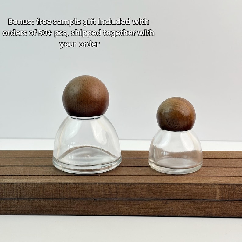 May include: Two clear glass bottles with wooden ball stoppers sit on a brown wooden surface. The bottles are different sizes. The text "Bonus: free sample gift included with orders of 50+ pcs, shipped together with your order" is in the background.