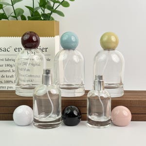May include: Clear glass perfume bottles with spherical stoppers in burgundy, blue, and yellow. Additional stoppers in white, black, and pink are also present. The bottles have silver spray nozzles.