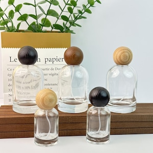 Puede incluir: Cinco frascos de perfume de vidrio transparente con tapones de madera en forma de bola. Los tapones son de varios tonos de marrón y beige. Los frascos están dispuestos sobre una superficie de madera, con una bolsa de papel y vegetación en el fondo.
