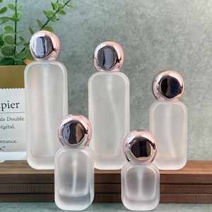 May include: Five frosted glass perfume bottles with rose gold caps. The bottles are rectangular with rounded corners. The caps are a shiny rose gold color. The bottles are arranged on a wooden surface.