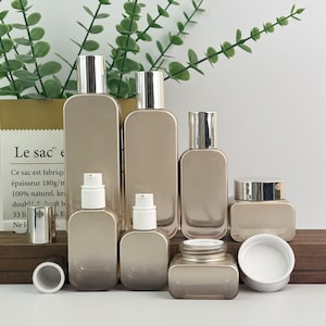 Peut inclure: Ensemble de flacons et pots cosmétiques dorés de différentes tailles, de forme rectangulaire, avec des bouchons argentés et des pompes blanches. Les contenants sont disposés sur une surface en bois, avec un sac en papier et de la verdure en arrière-plan.