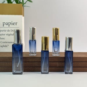 May include: Five small, rectangular glass perfume bottles with silver and gold caps. The bottles have a blue gradient color and are arranged on a wooden surface. The background includes a paper bag with French text.