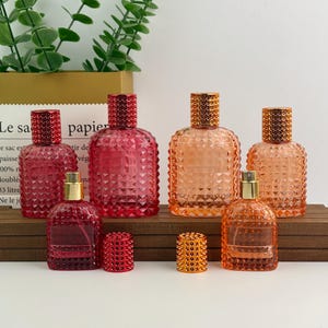 May include: An assortment of decorative perfume bottles in shades of red and orange. The bottles have textured, diamond-shaped patterns and are topped with studded caps. Some bottles have gold-colored sprayers.