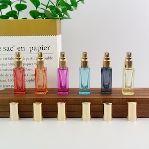 May include: Six rectangular glass perfume bottles with gold spray nozzles, arranged in a row. The bottles are red, orange, pink, blue, dark gray, and yellow. Six matching gold caps are placed in a line in front of the bottles on a wooden surface.