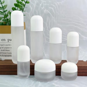 Puede incluir: Un conjunto de envases cosméticos de vidrio esmerilado con tapas blancas redondeadas. El conjunto incluye botellas y tarros de varios tamaños, adecuados para lociones, sueros o cremas. Los envases están dispuestos sobre una superficie de madera.