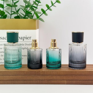 Può includere: Quattro flaconi di profumo in vetro con scanalature verticali e ugelli spray dorati. Due flaconi presentano una finitura sfumata verde con tappi verde scuro, mentre gli altri due hanno una sfumatura grigia con tappi neri, disposti in fila.