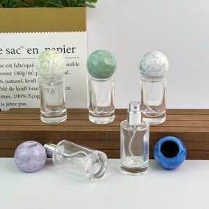 Könnte beinhalten: Klare Parfümflaschen aus Glas mit dekorativen kugelförmigen Stöpseln in verschiedenen Farben, darunter Grün, Weiß und Blau. Eine Flasche liegt auf der Seite. Die Flaschen werden auf einer Holzoberfläche präsentiert.