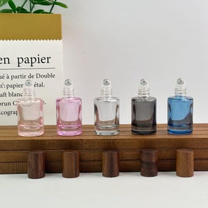 May include: Five small glass roller bottles with metal ball tops, arranged in a row. The bottles are clear, light pink, clear, dark gray, and blue. Five cylindrical dark wood caps are placed in a line directly in front of the bottles.