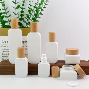 Peut inclure: Une collection de contenants cosmétiques blancs aux formes rectangulaires arrondies et lisses, dotés de bouchons en bois clair. L'ensemble comprend des bouteilles de tailles variées avec pompes et bouchons à vis, ainsi que des pots carrés assortis.