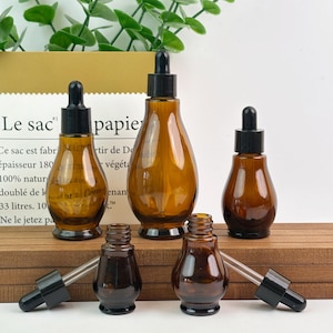 Könnte beinhalten: Eine Auswahl an braunen Glas-Tropfflaschen mit schwarzen Kappen und Pipetten. Die Flaschen haben unterschiedliche Größen und Formen, darunter ein Tropfendesign. Im Hintergrund befindet sich eine Papiertüte mit französischem Text.