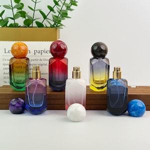 May include: A collection of colorful perfume bottles with spherical stoppers. The bottles are various colors, including green, red, purple, white, yellow, and blue. Each bottle has a gold-colored atomizer. Decorative spheres in matching colors are in front of the bottles.