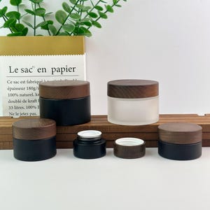 Könnte beinhalten: Ein Set aus Kosmetikdöschen mit dunkelbraunen Holzmaserungsdeckeln. Es umfasst zwei große mattschwarze Tiegel, einen großen Tiegel aus Milchglas, einen kleinen mattschwarzen Tiegel mit weißem Inneneinsatz und einen einzelnen Holzdeckel.