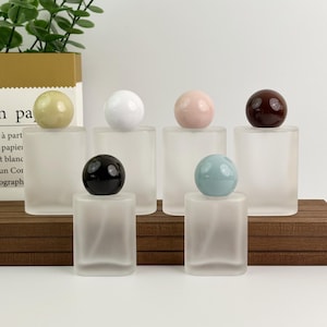 Puede incluir: Seis frascos de perfume de vidrio esmerilado con tapones esféricos en varios colores: verde, blanco, rosa, marrón oscuro, negro y azul. Los frascos son rectangulares y se apoyan sobre una superficie de madera, mostrando un diseño minimalista.