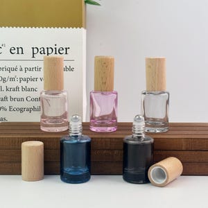 May include: Five small glass roller bottles with light wood caps. Three bottles stand upright with caps on, featuring clear, pink, and clear glass. Two bottles sit in front with metal roller balls, one blue and one black, with a detached wood cap nearby.